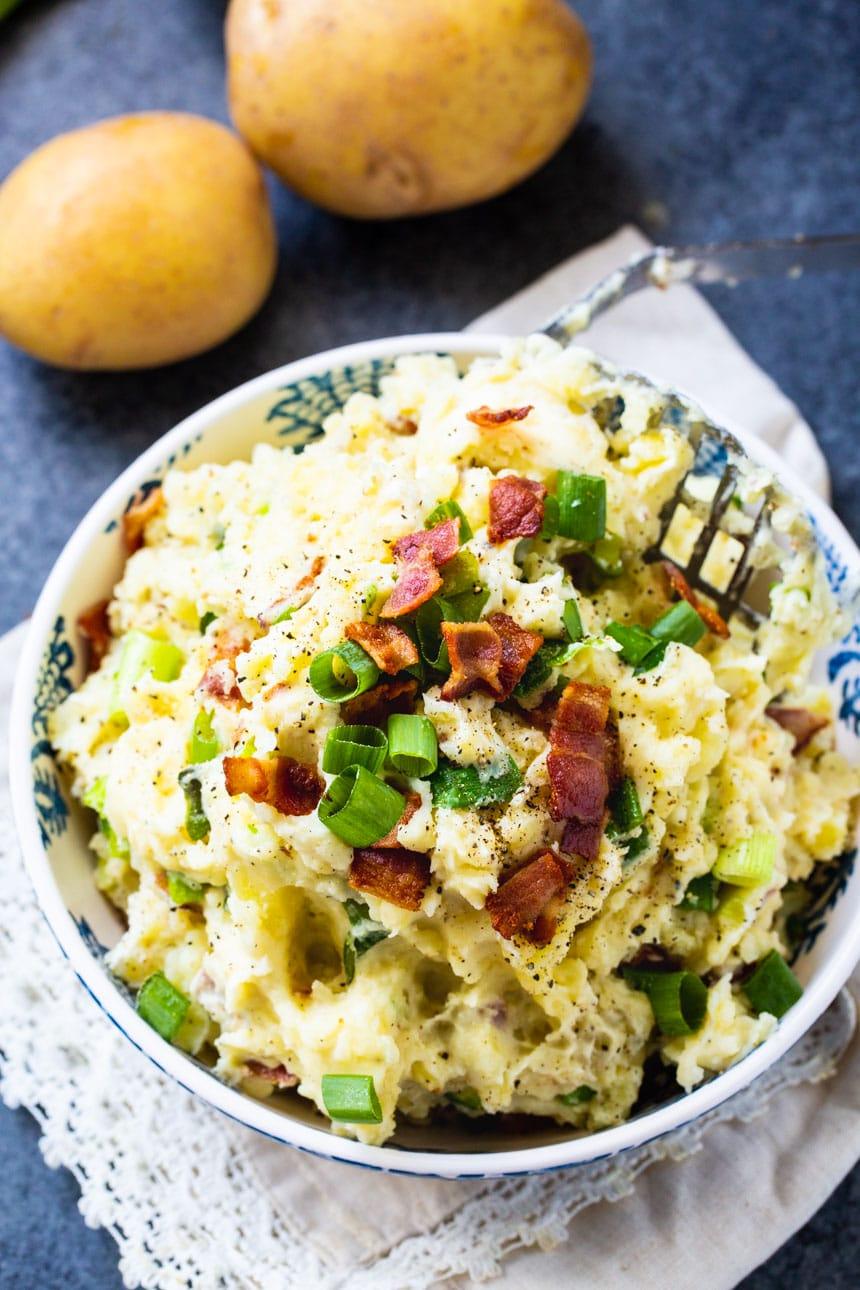 Overhead of Bacon-Green Onion Mashed Potatoes in a bowl with potatoes beside the bowl.