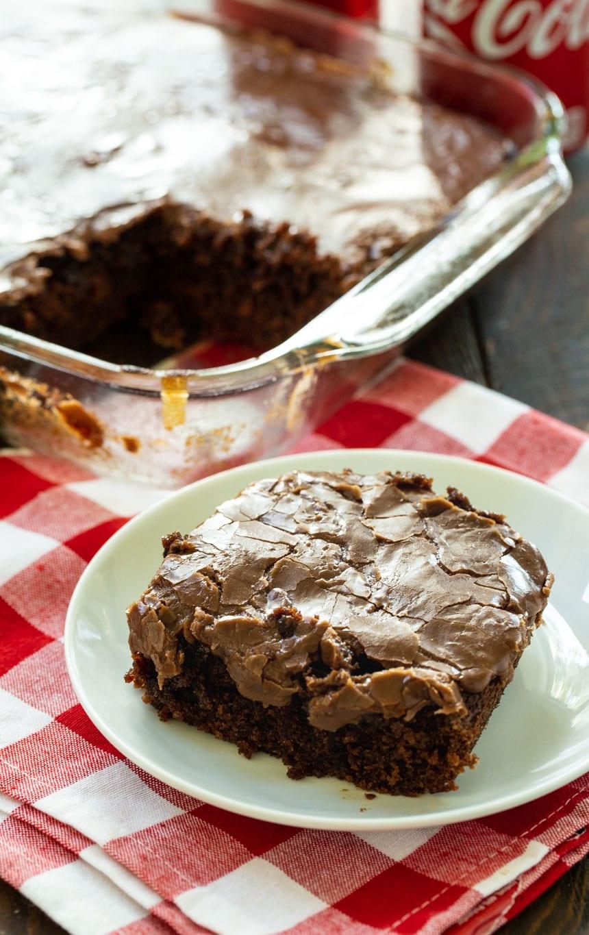 Slice of Coca Cola Cake on a white plate with remaining cake in the background.
