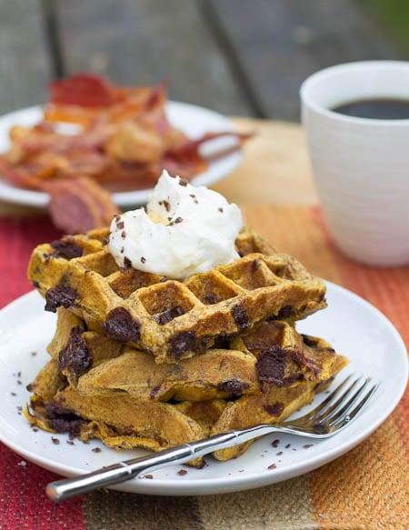 Three Pumpkin Waffles stacked on a plate with bacon in background.