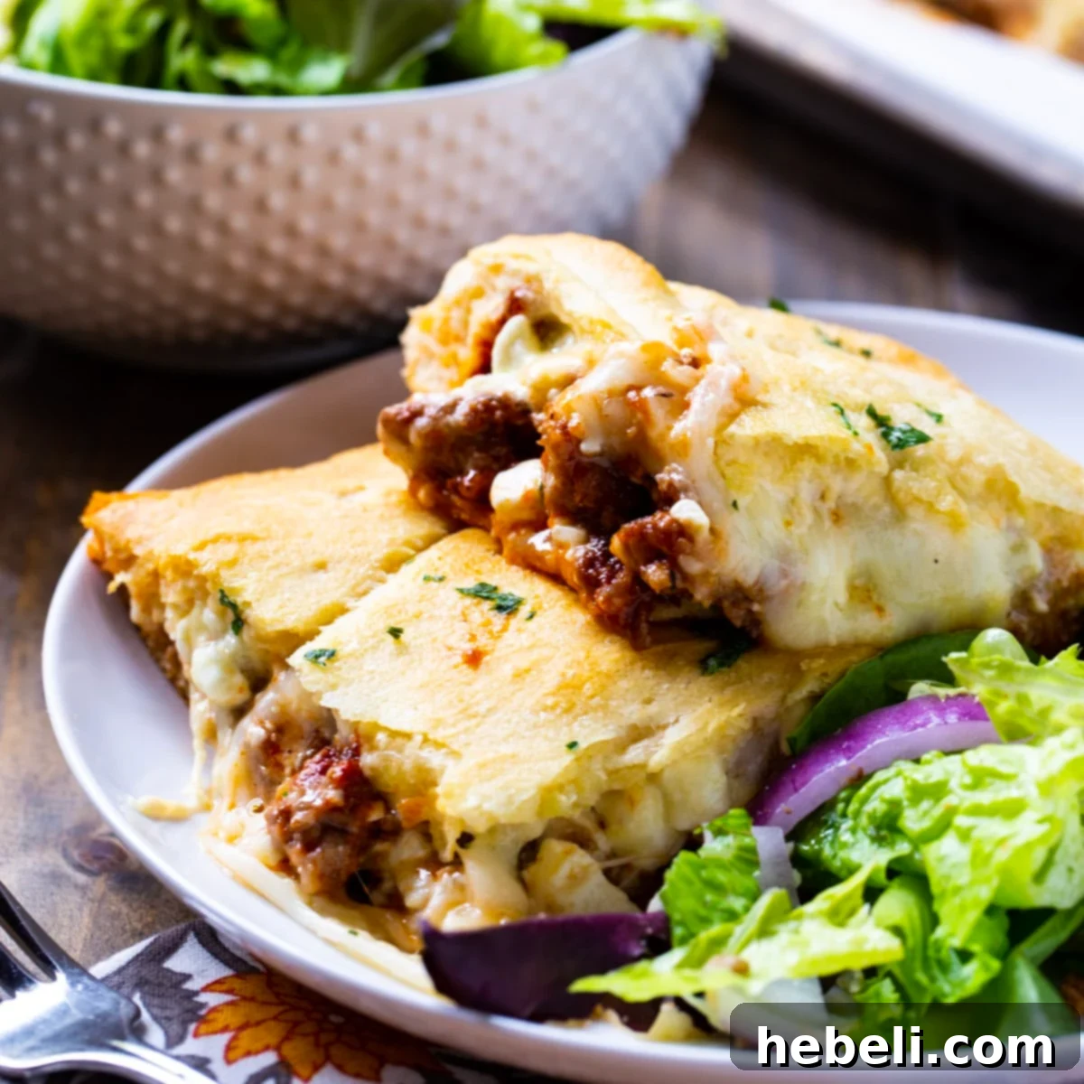 A serving of golden-baked Crescent Lasagna on a plate with a fresh green salad, ready to enjoy. The flaky crust and delicious layers are visible.