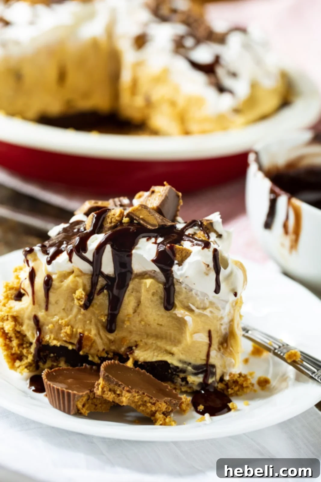 A close-up view of a slice of Peanut Butter Pie on a plate, with the rest of the pie blurred in the background, clearly showing the distinct layers of graham cracker crust, chocolate, and the creamy peanut butter filling topped with whipped cream and chocolate drizzle.