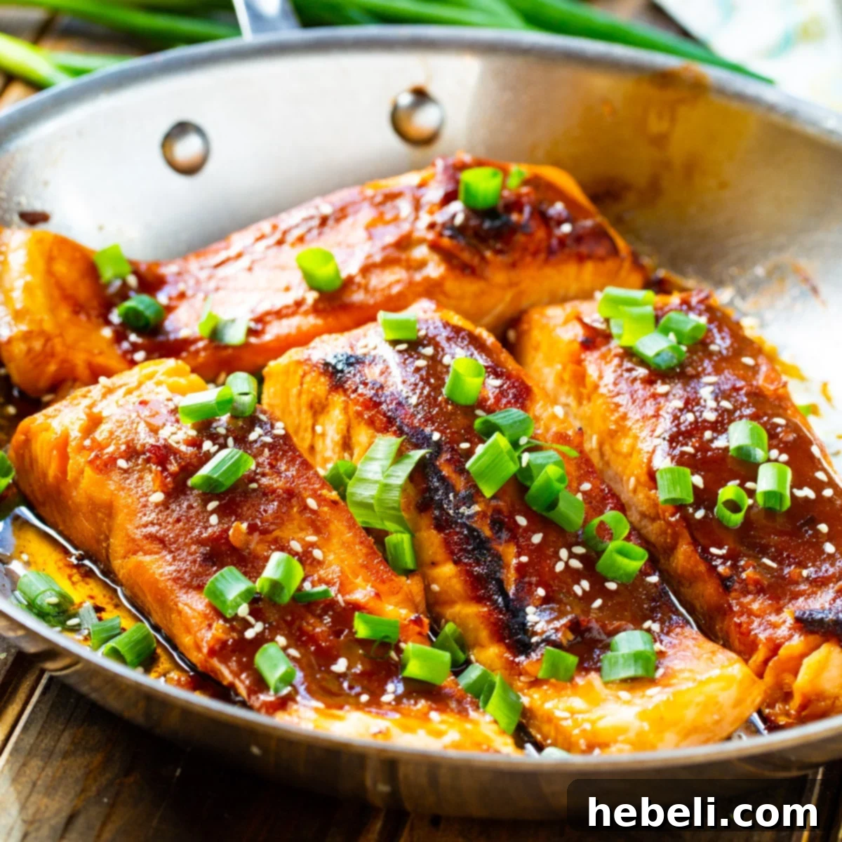 Four golden-brown Teriyaki Salmon fillets resting in a cast iron pan, garnished with green onions and sesame seeds.