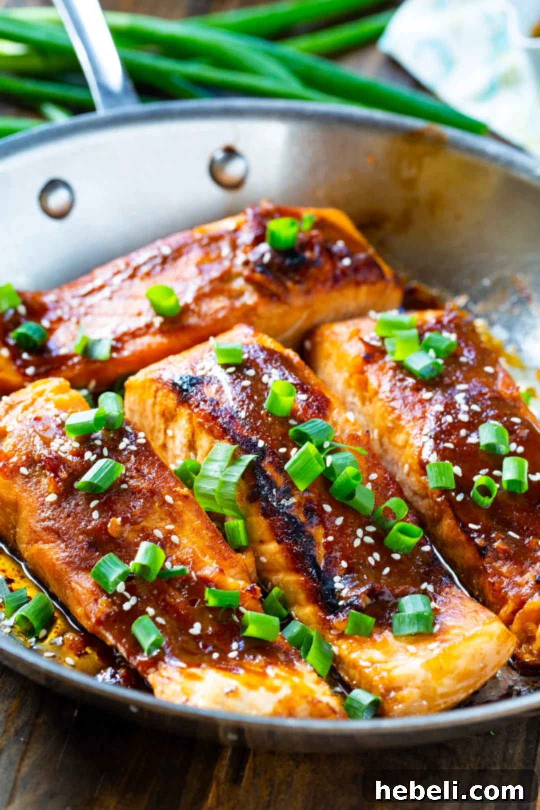 Two pieces of cooked teriyaki salmon glistening in a stainless steel pan, garnished with sliced green onions.