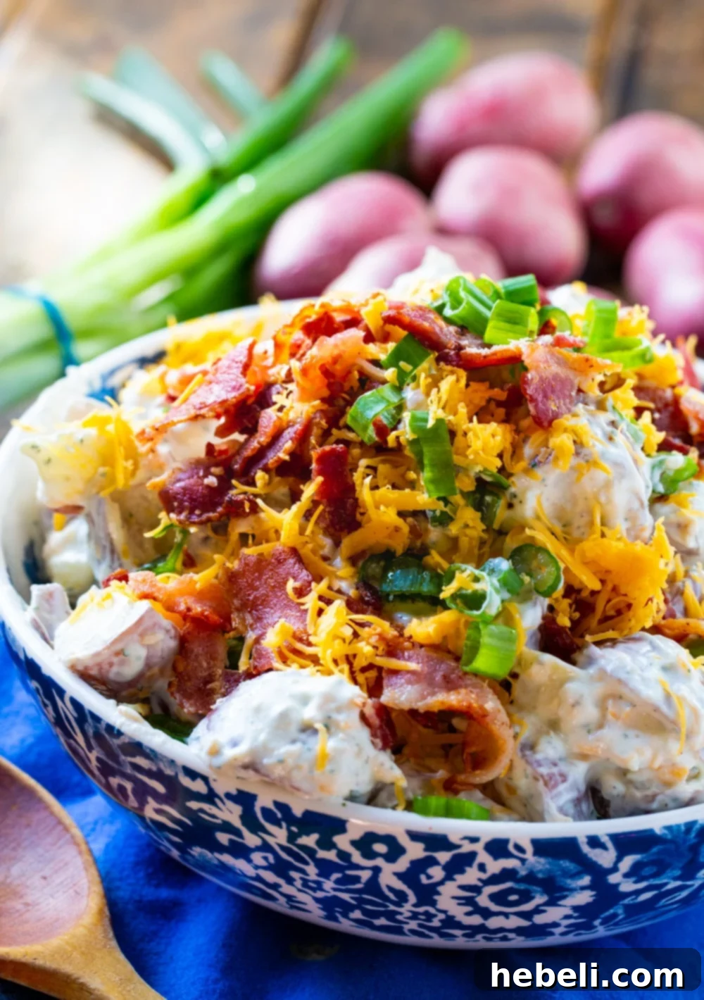 Creamy potato salad presented in a decorative bowl, with a blurred background showing whole cooked potatoes and chopped green onions, highlighting the salad's appealing texture.