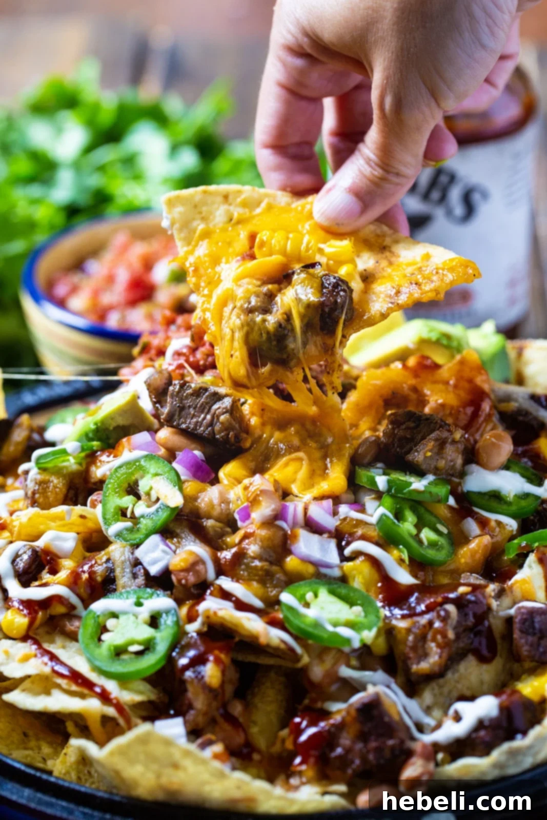 A hand reaching for a perfectly loaded BBQ Steak Nacho, showcasing the blend of grilled rib-eye, pinto beans, and corn.