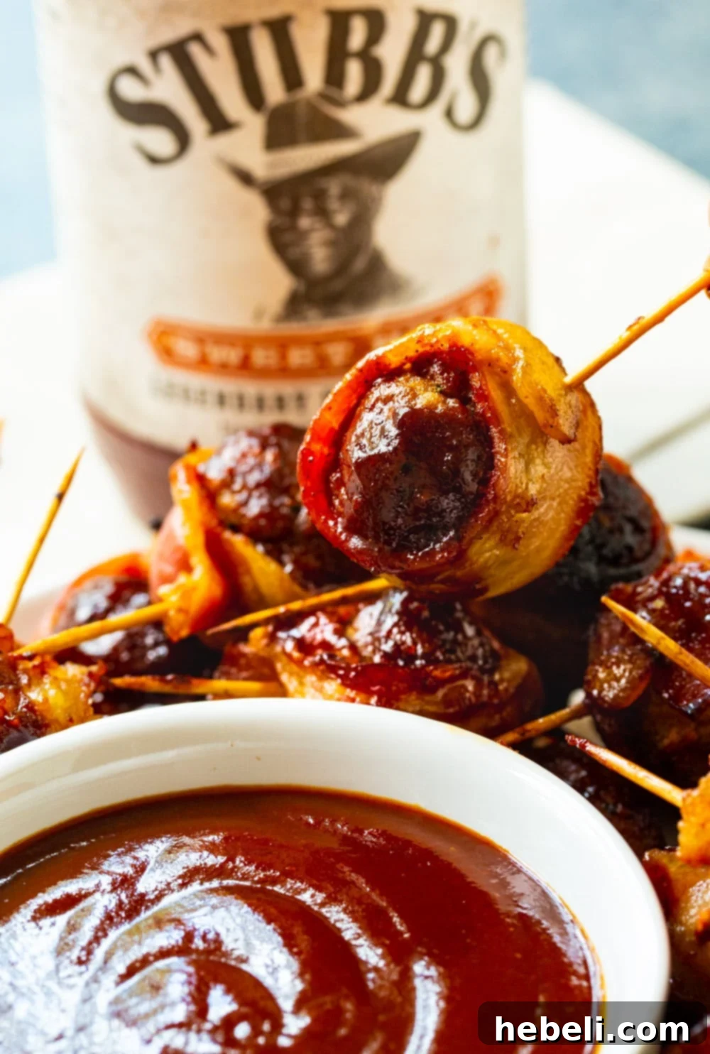 A Barbecue Bacon Meatball being lifted by a toothpick, ready to be dipped into a small bowl of savory barbecue sauce.