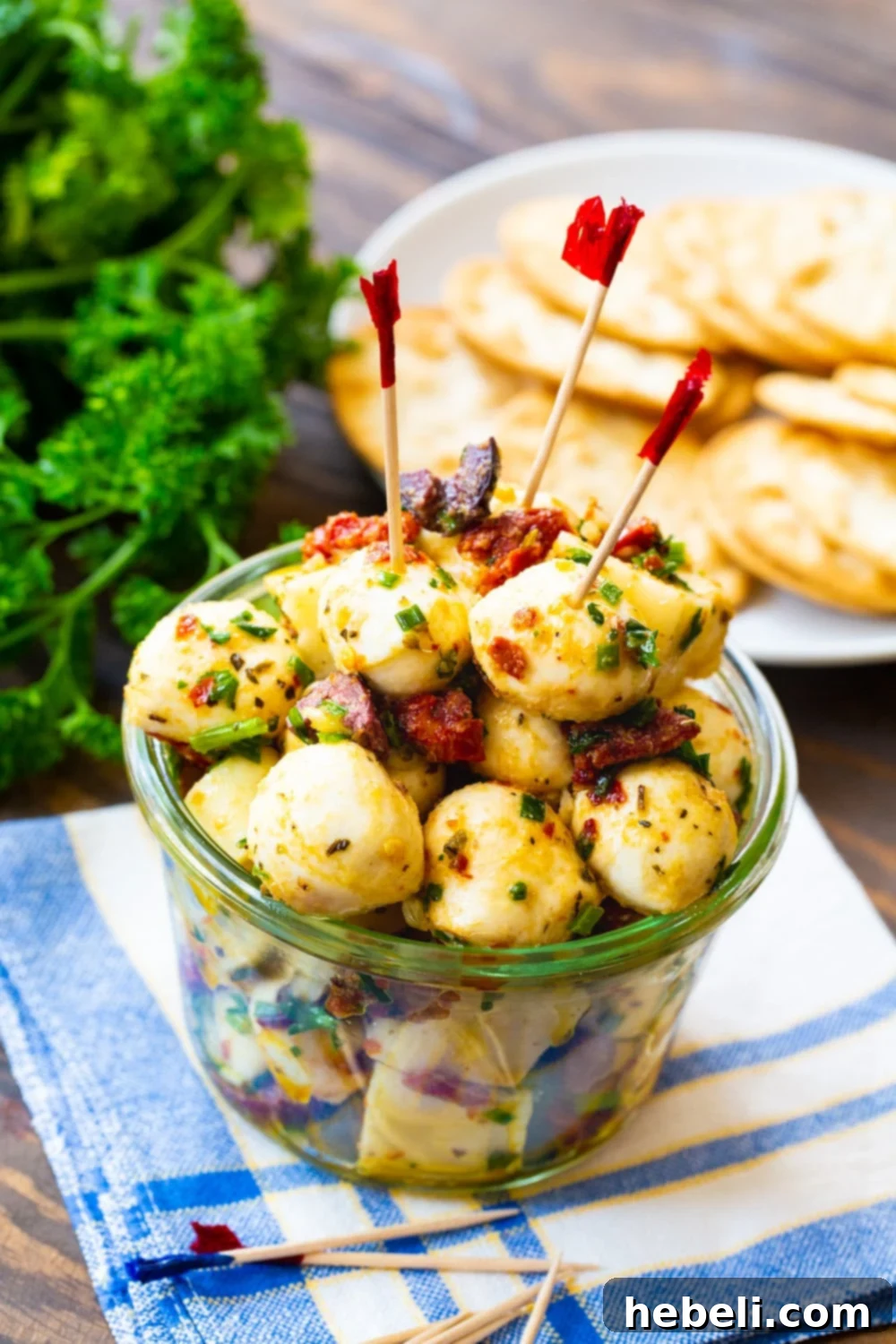 Glass jar filled with spicy marinated mozzarella balls, flanked by crackers and fresh parsley.