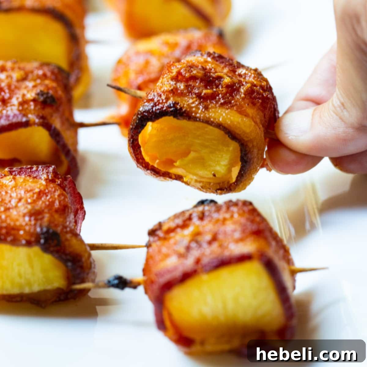 Hand picking up a perfectly cooked Spicy Bacon Wrapped Pineapple Bite, showcasing the crispy bacon and juicy pineapple.