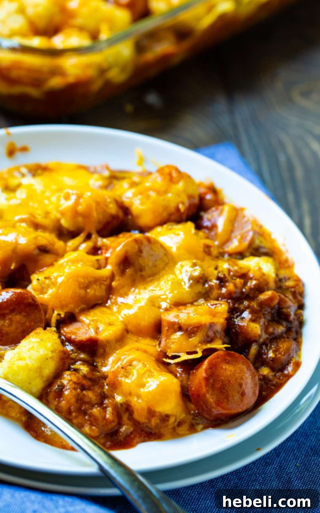 A portion of the casserole on a white plate, showing melted cheese, tater tots, and chili hot dogs.