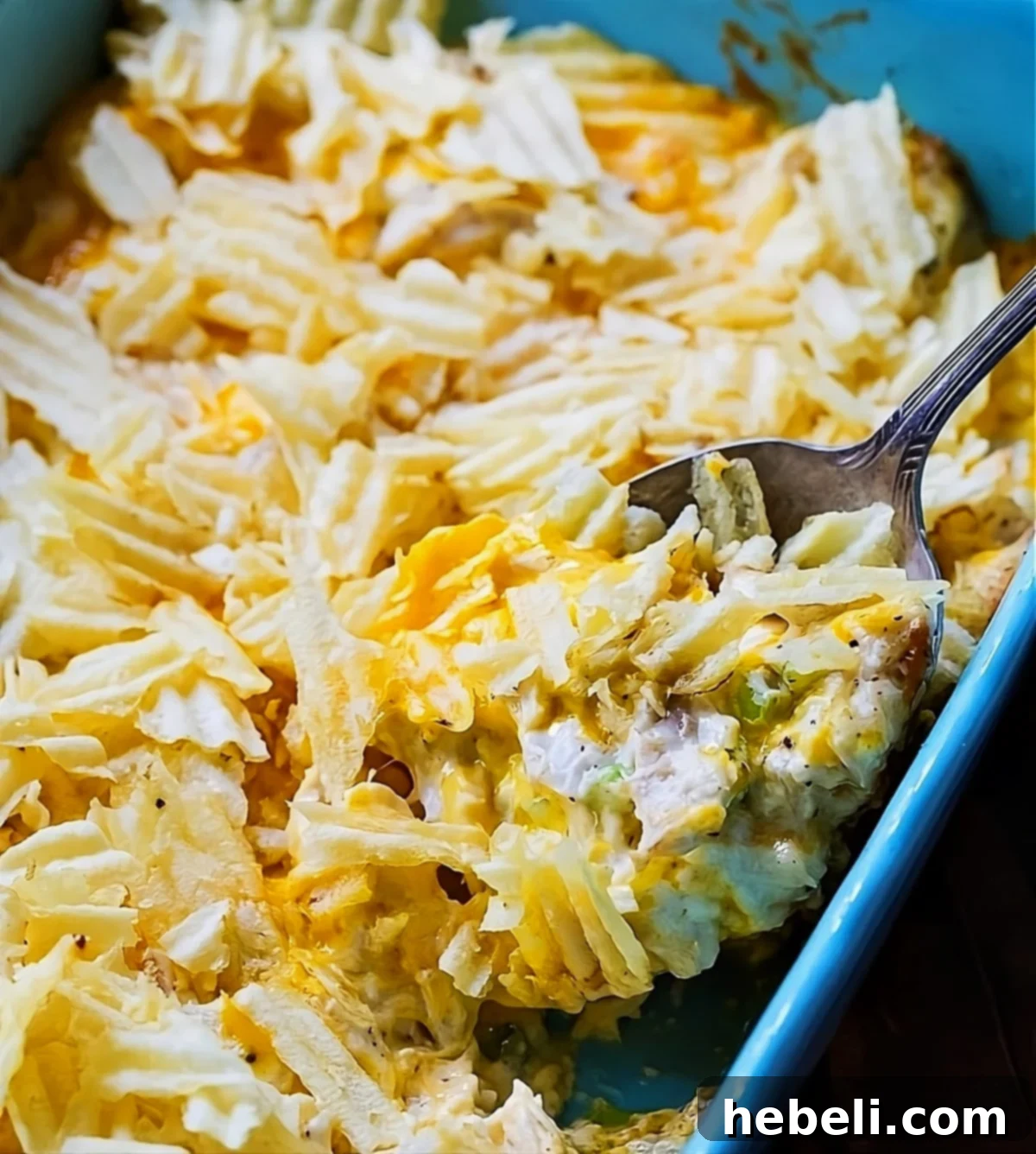 Warm Chicken Salad Bake 2 A spoon scooping a generous portion of Hot Chicken Salad from a casserole dish, revealing its creamy texture, chunks of chicken, and golden-brown, crispy potato chip topping.
