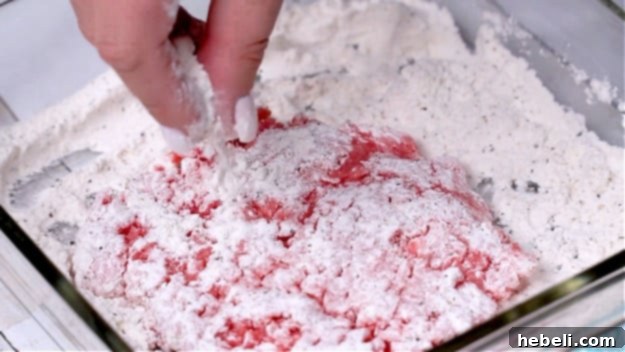 Smothered Steak Bites with Golden Onion Gravy 5 A hand pressing a piece of cubed steak into a flour mixture in a shallow dish, ensuring it's evenly coated.