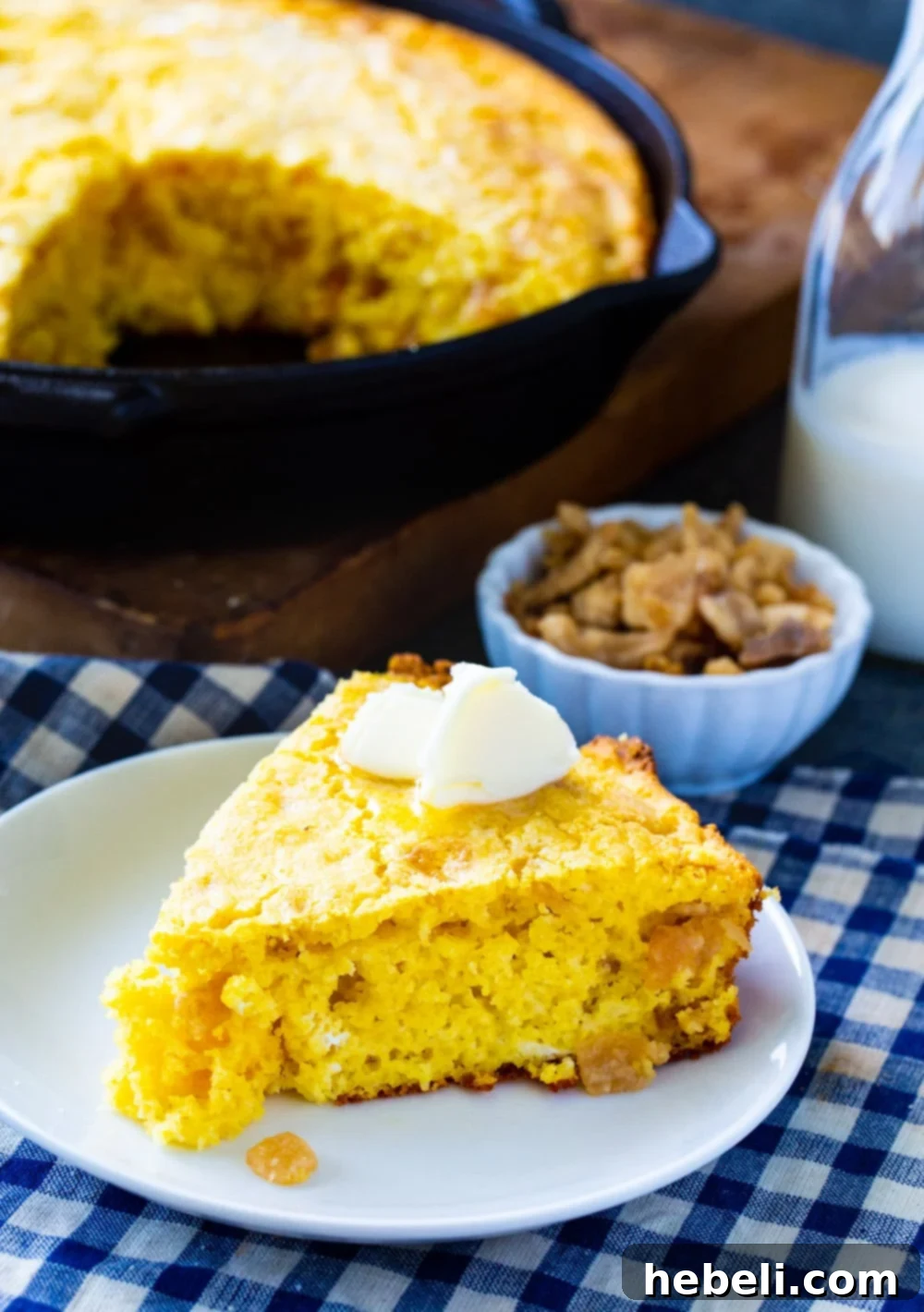 A delicious slice of golden-brown cornbread, showing its tender crumb and crackling bits, served on a plate with the cast iron pan full of the remaining cornbread in the background.