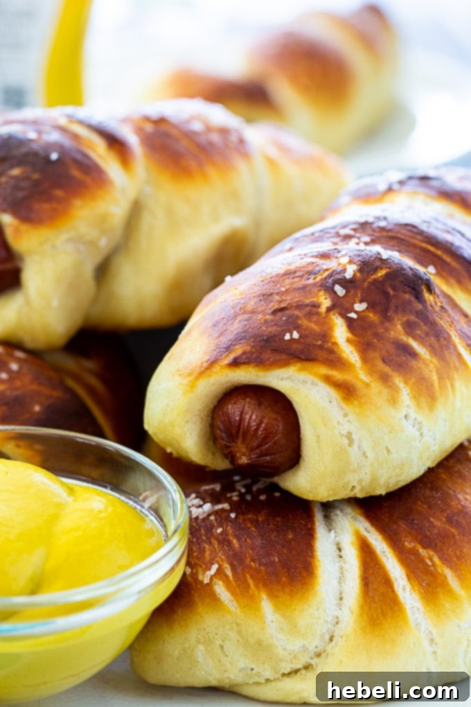 A close-up of a single homemade Pretzel Dog, beautifully golden brown and generously sprinkled with coarse salt, sitting on a white background.