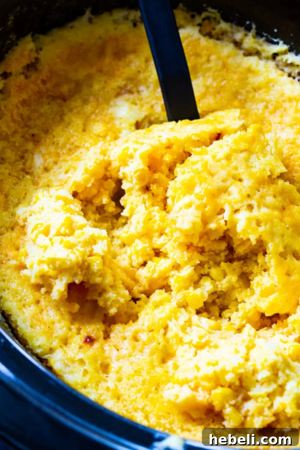 A serving spoon lifting a scoop of golden, bubbling Crock Pot Corn Casserole directly from the slow cooker, showing its rich, cheesy interior.