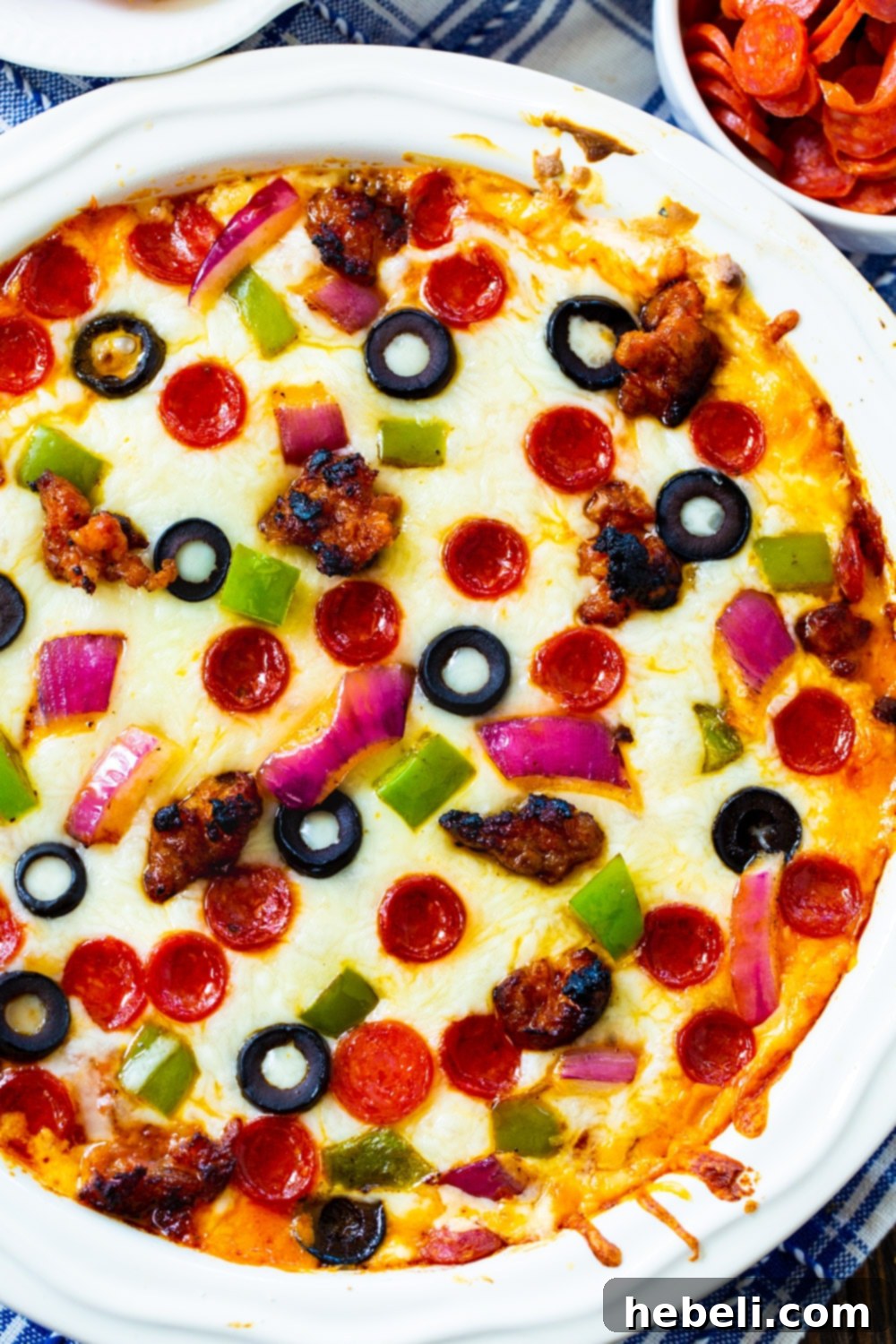 A white pie plate filled with the layered Supreme Pizza Dip before baking, showing raw sausage, pepperoni, onion, and bell pepper on top of sauce and cheese.