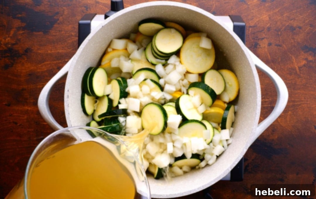 Chicken broth being poured into a pot containing sliced squash, zucchini, and chopped onion, ready for simmering.