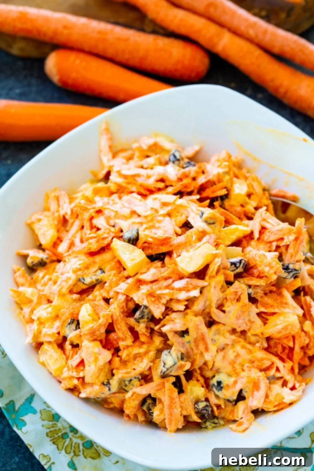 Sunshine Carrot Raisin Salad 3 Close-up of Carrot Raisin Salad in a rustic bowl, surrounded by fresh, vibrant carrots on a wooden surface.