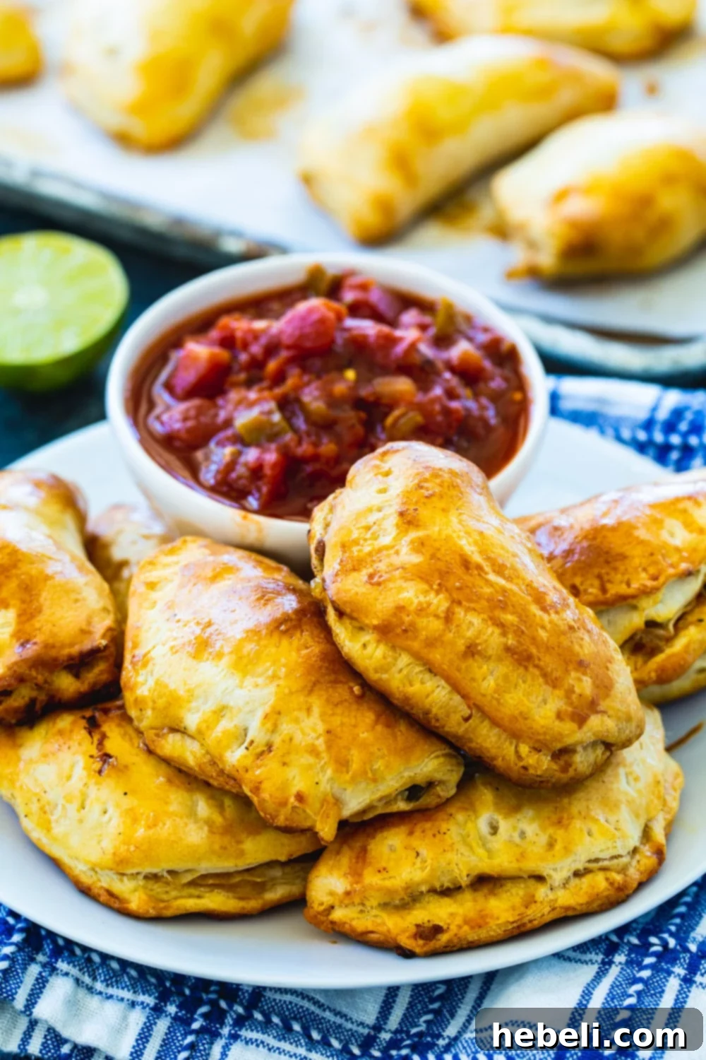 A plate of freshly baked Empanadas, perfectly golden, with a bowl of salsa. More empanadas are visible on a baking sheet in the background.