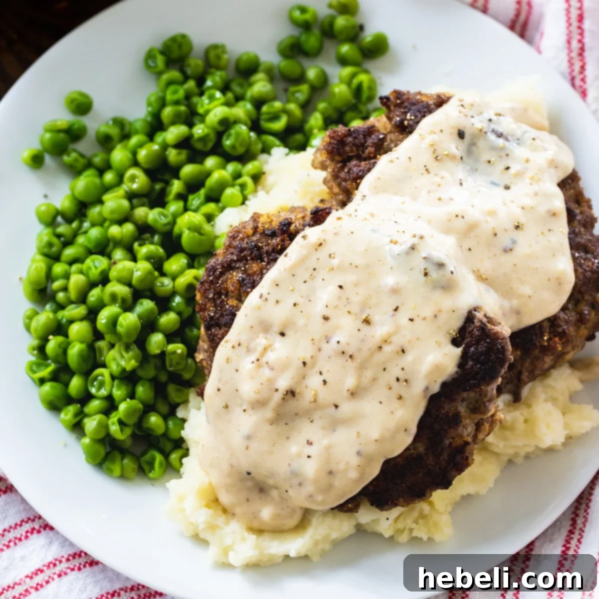 Classic Hamburger Steaks with creamy country gravy served over a bed of fluffy mashed potatoes, accompanied by vibrant green peas, creating a comforting and delicious plate.
