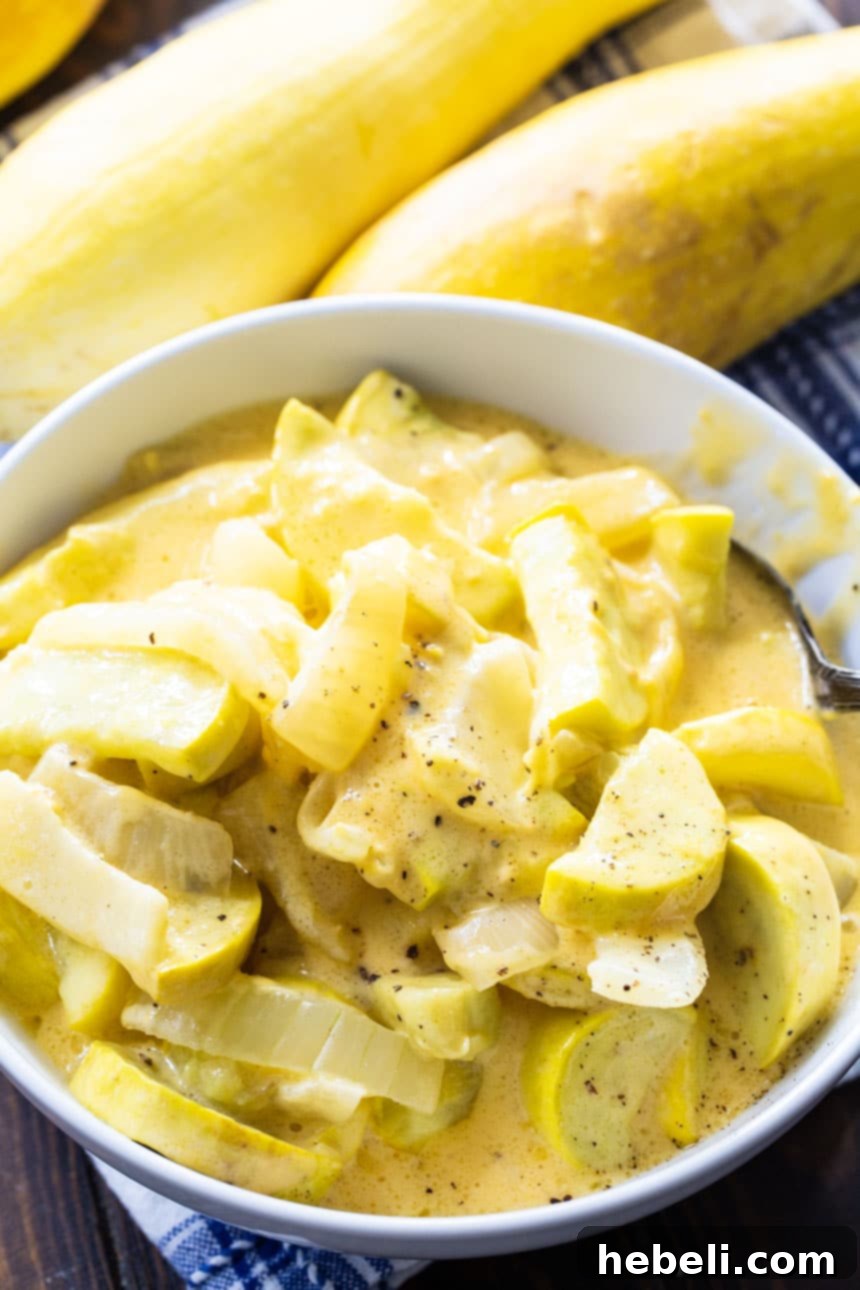 Cheesy Yellow Squash served in a white bowl, with fresh yellow squash and herbs in the background.