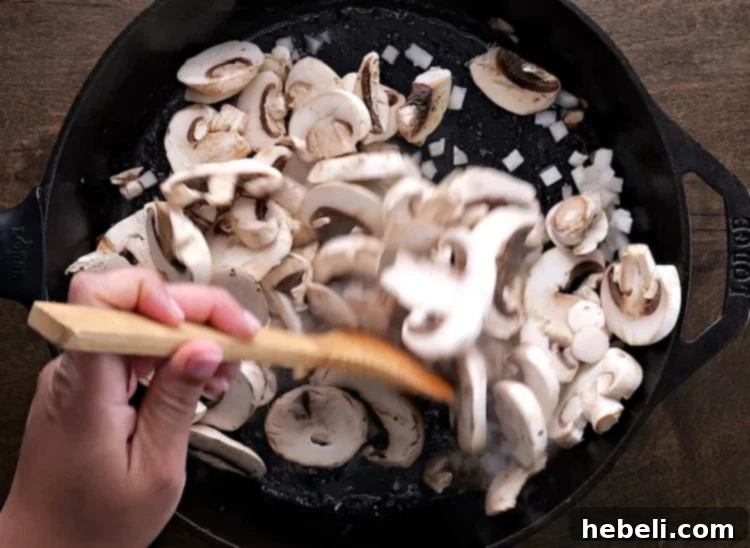 Sliced cremini mushrooms and diced onions actively sautéing in a skillet with butter and pan drippings, turning golden and fragrant.