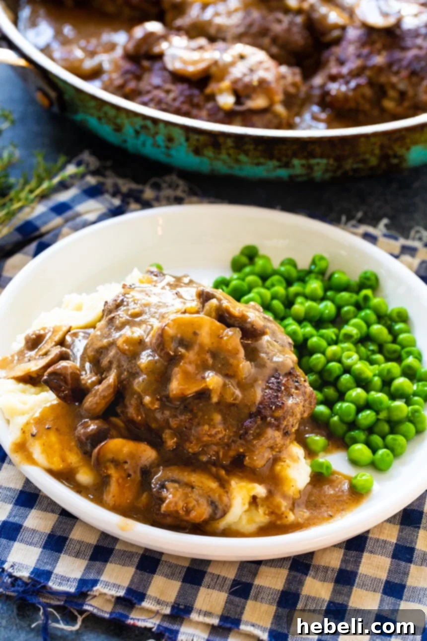 A close-up shot of a single Hamburger Steak generously covered in rich, thick mushroom gravy, served on a white plate alongside a portion of green peas, inviting a delicious meal.
