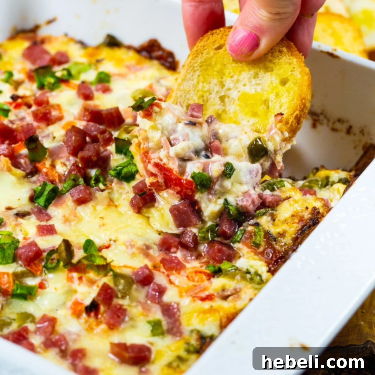 A toasted baguette slice generously scooping up creamy Hot Muffaletta Dip, rich with olives and meats.