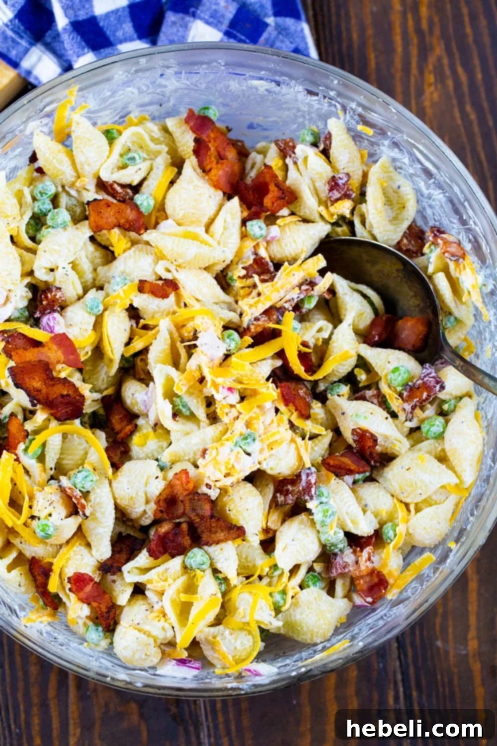 A close-up view of creamy pasta salad being gently mixed in a large stainless steel bowl, showcasing the consistency of the dressing and the fresh inclusions.