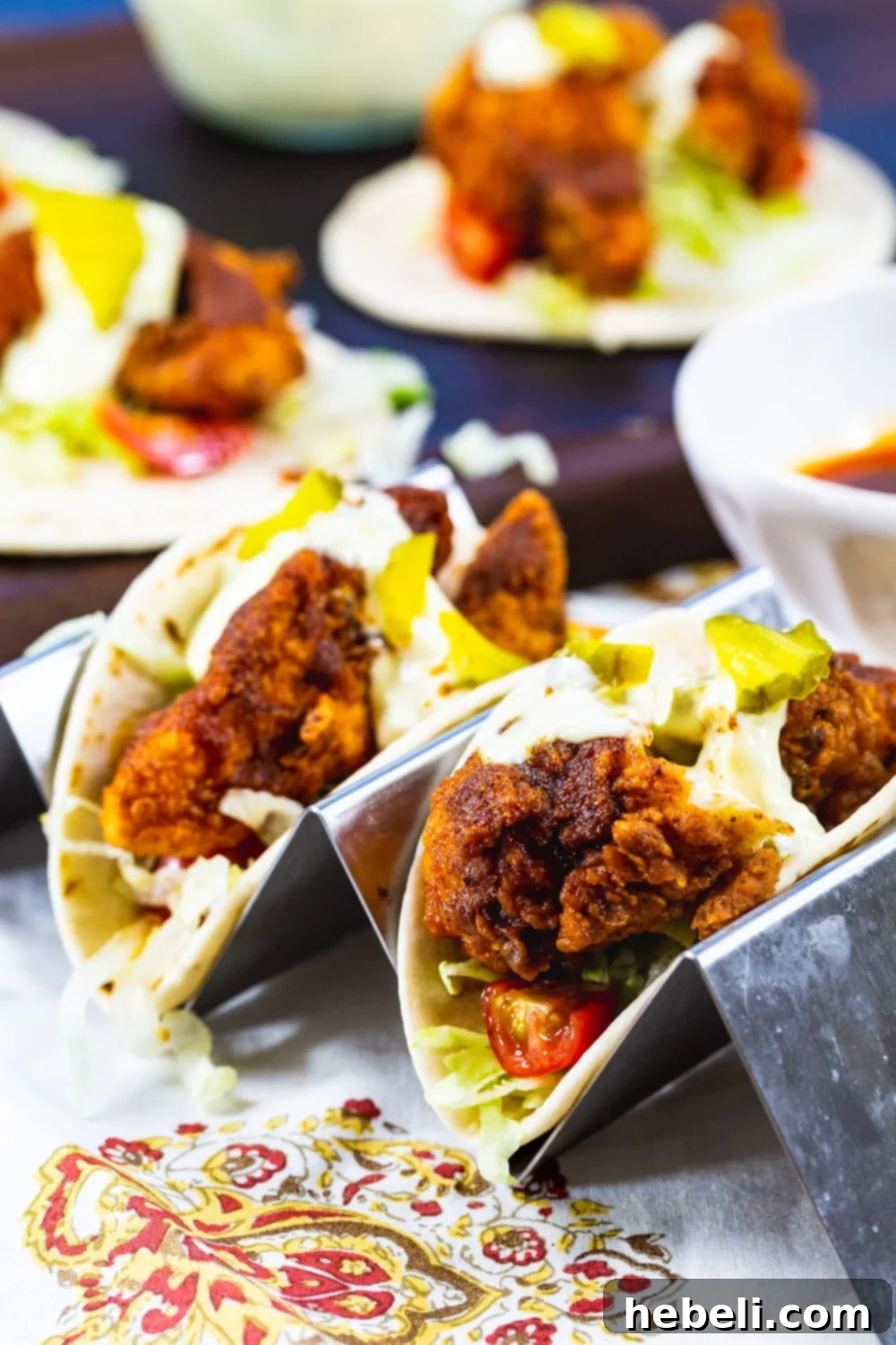Blazing Nashville Chicken Tacos 4 Two Nashville Hot Chicken Tacos presented in a tray, with more tacos blurred in the background.