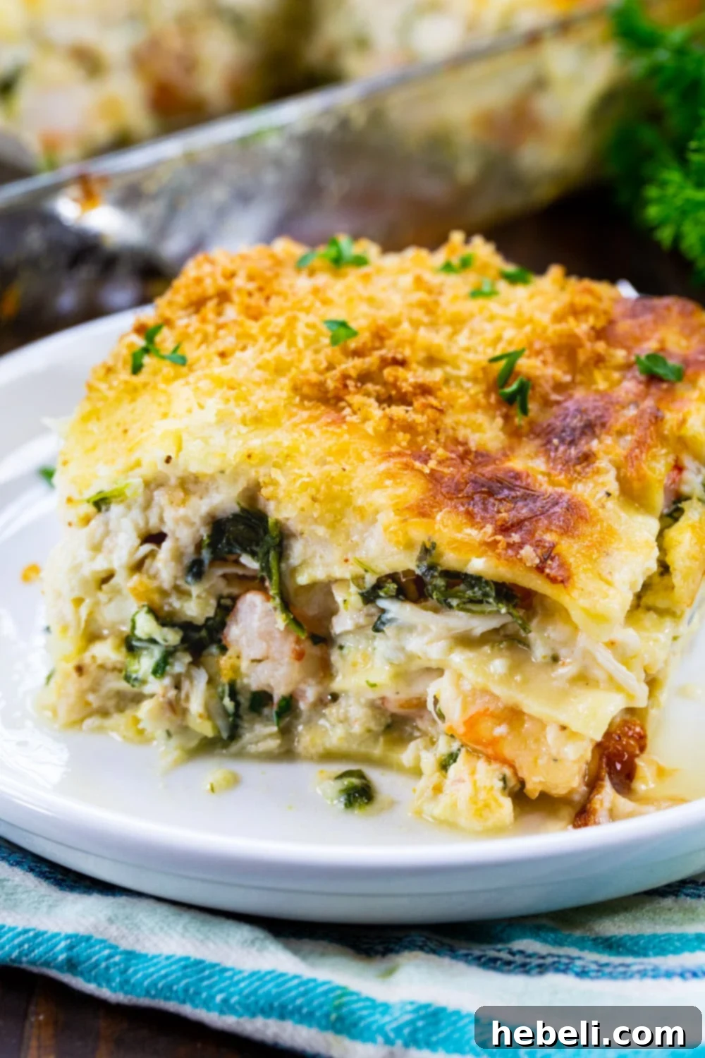 A close-up of a slice of White Seafood Lasagna on a plate, showing the creamy white sauce, noodles, shrimp, crab, scallops, and green spinach.