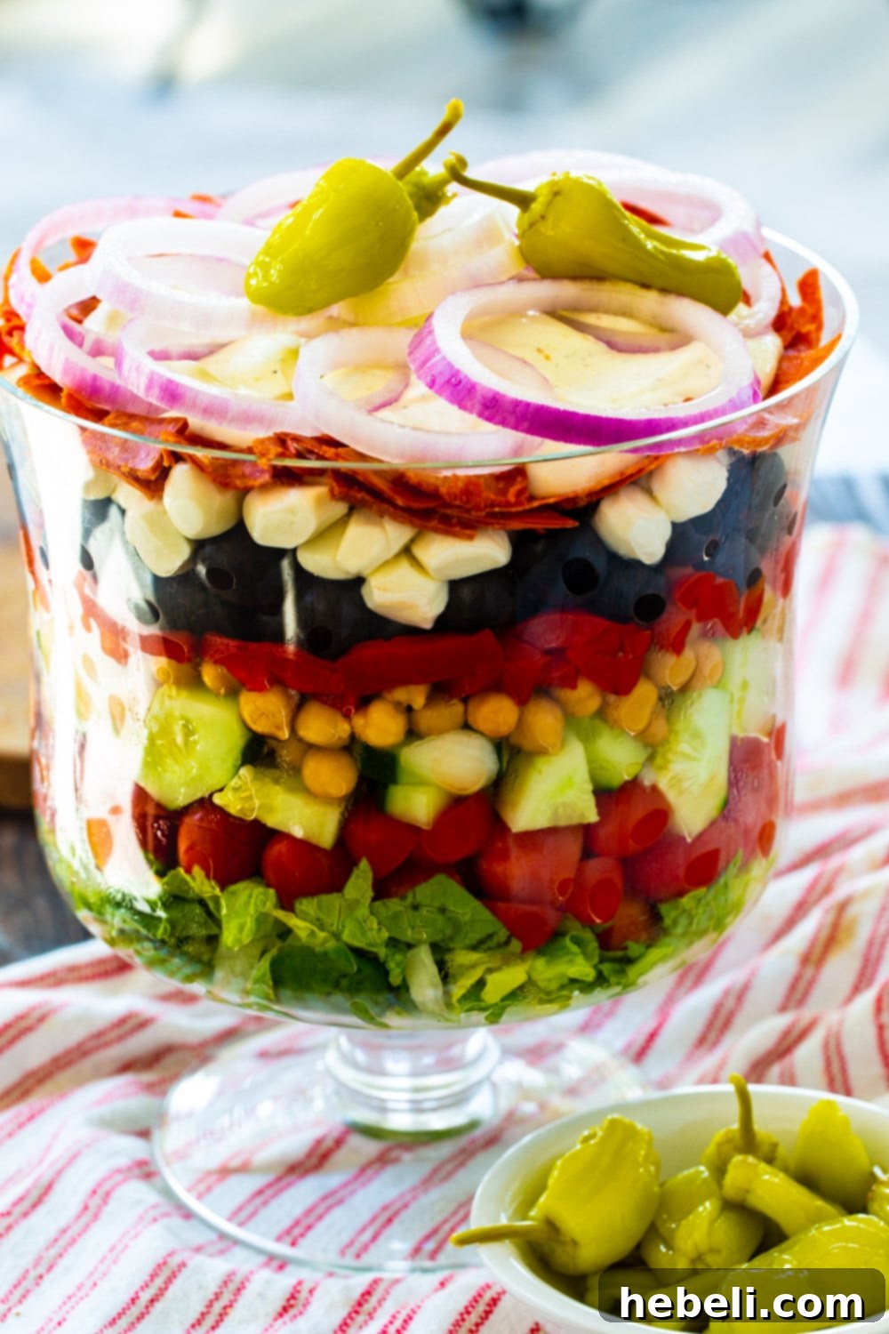 Italian Layered Salad elegantly presented in a glass trifle bowl, with a small bowl of tangy pepperoncini peppers placed in front.