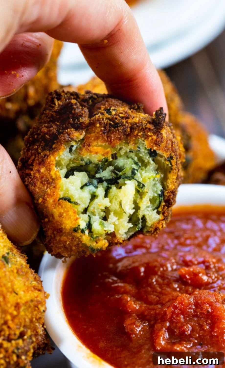 A hand dipping a golden crispy spinach cheese ball into a bowl of vibrant red pizza sauce.