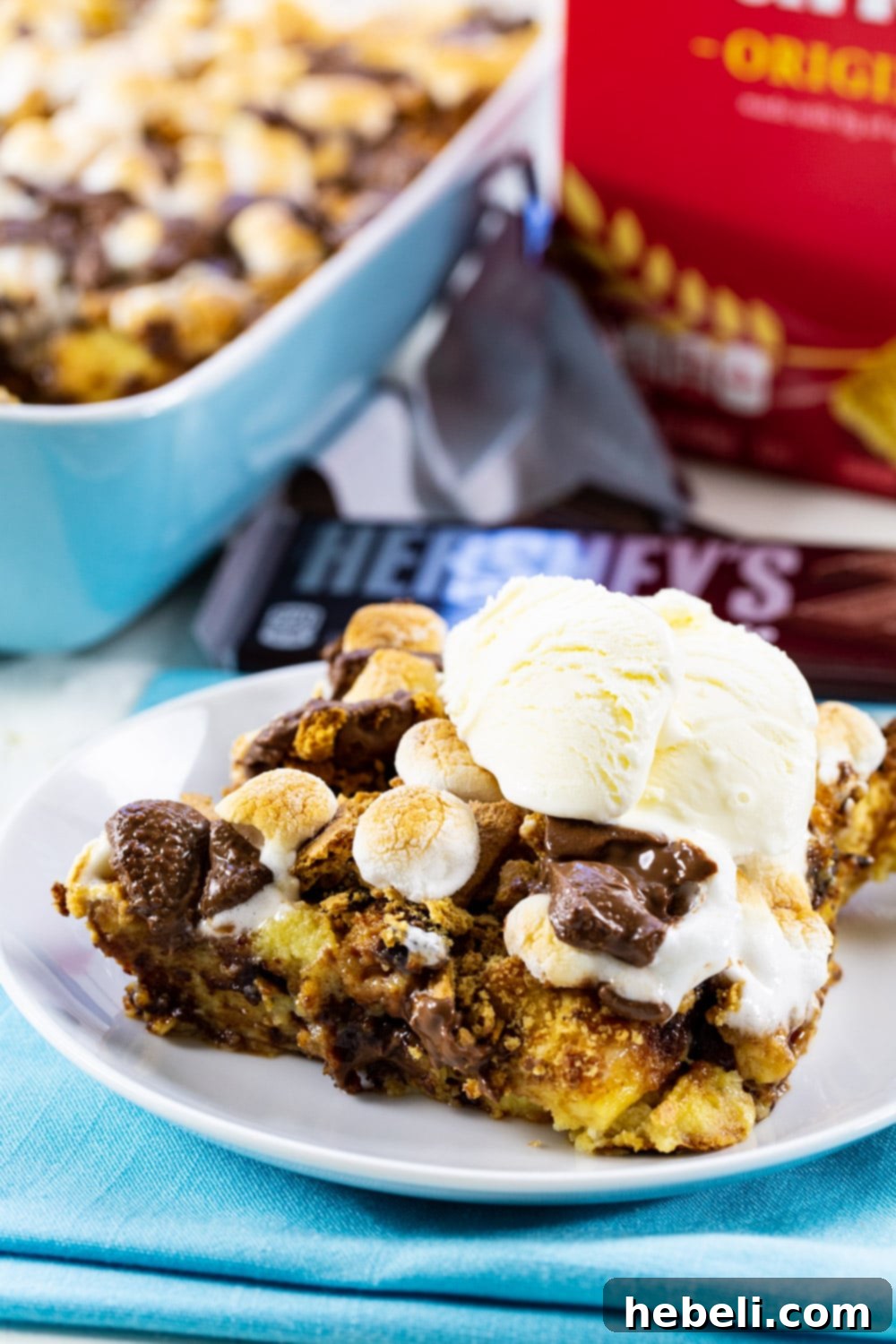 A generous serving of S'mores Bread Pudding, glistening with melted chocolate and marshmallows, crowned with a scoop of vanilla ice cream for a perfect contrast of warm and cold.
