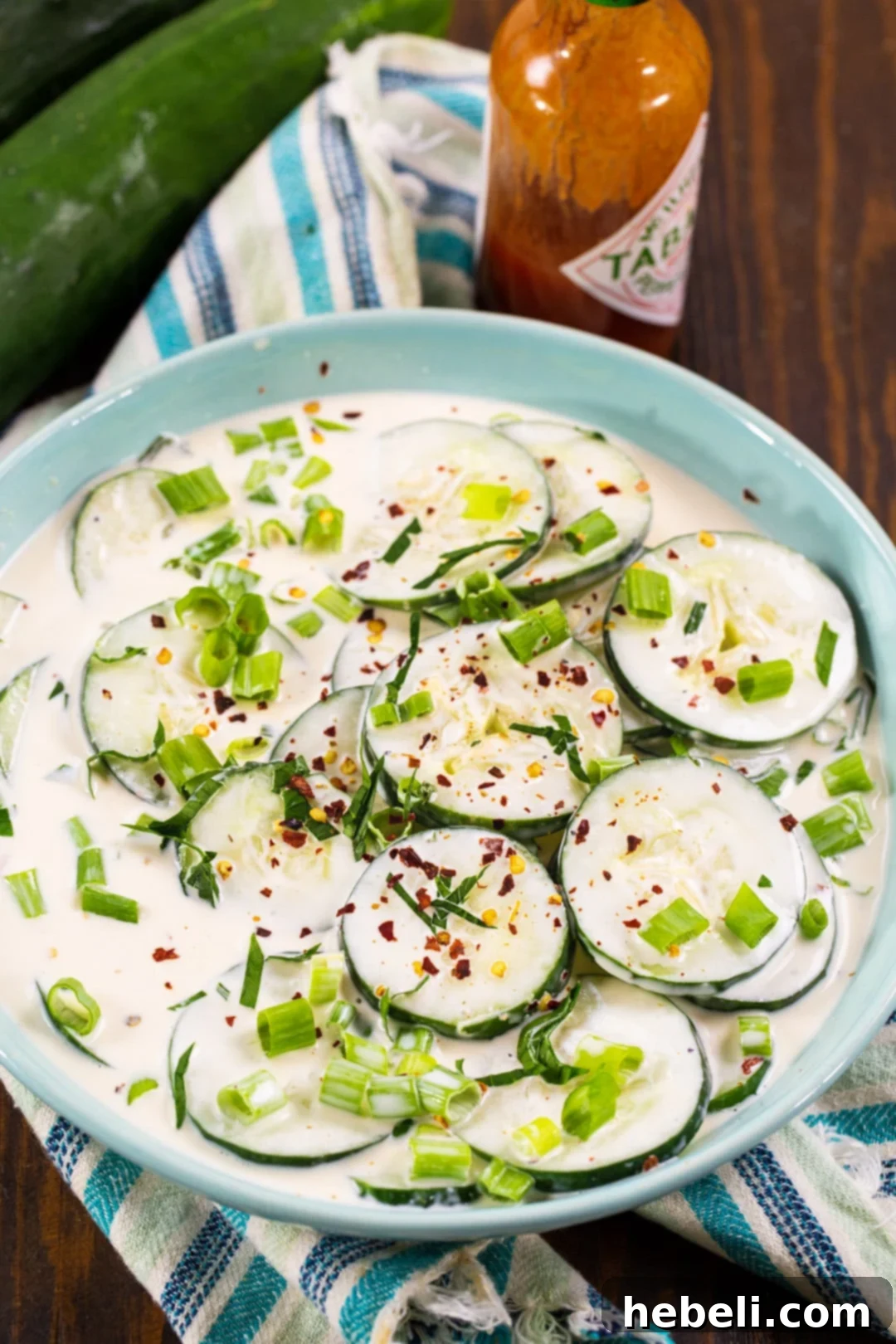 A close-up shot of Creamy Spicy Cucumbers in a blue ceramic bowl, with a bottle of hot sauce in the background, highlighting the vibrant colors and creamy texture.