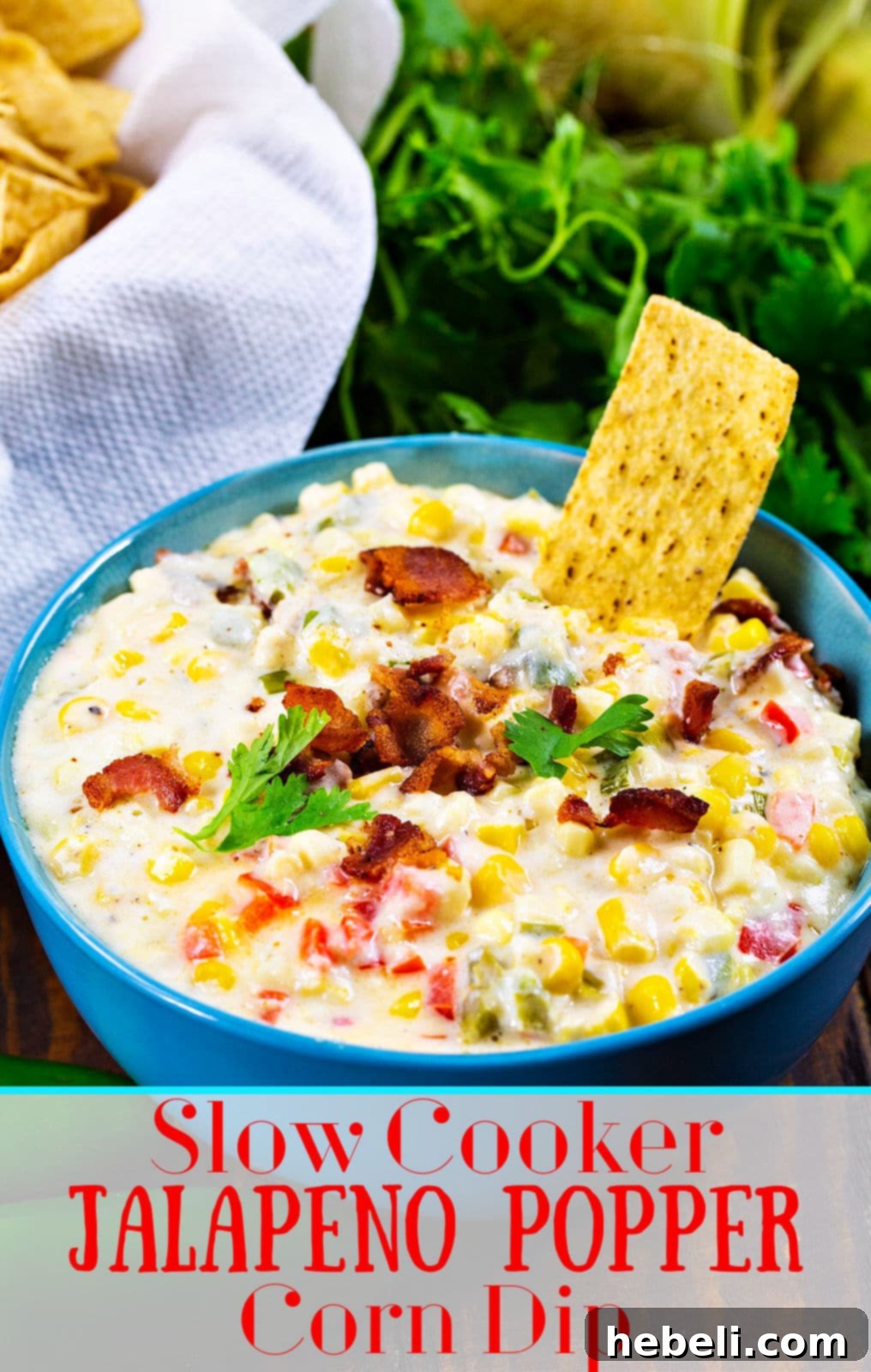 Zesty Cheesy Slow Cooker Corn Dip 5 A wide shot of a blue bowl filled with Slow Cooker Jalapeno Popper Corn Dip, beautifully garnished with fresh cilantro and crispy bacon, thoughtfully placed on a rustic wooden table with scattered tortilla chips.