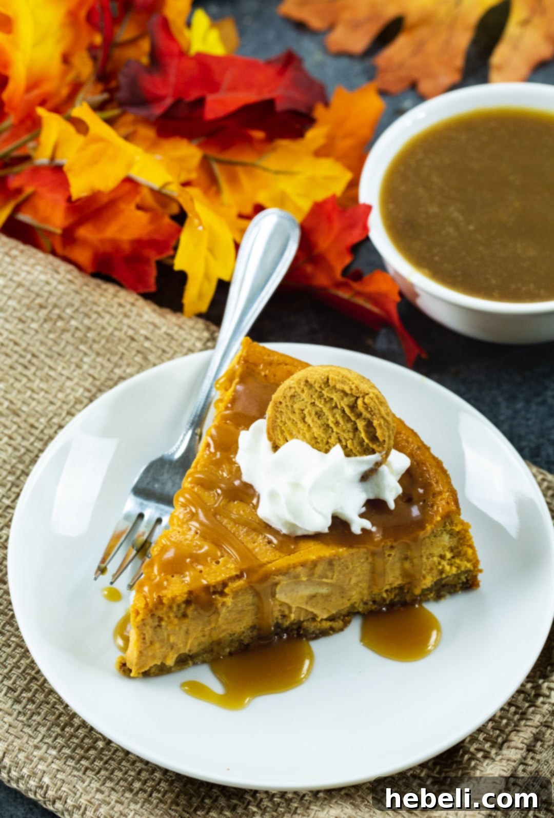 A complete Pumpkin Cheesecake with Gingersnap Crust, beautifully plated alongside scattered fall leaves and a small bowl of golden caramel sauce, evoking a cozy autumn scene.