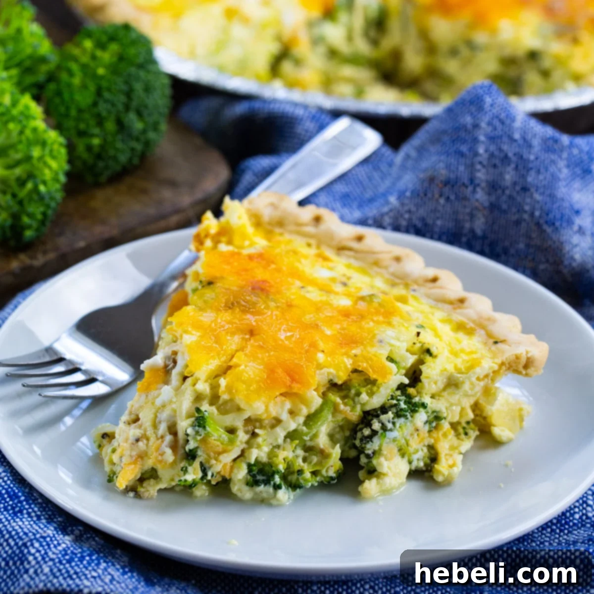A perfectly baked slice of golden-brown Broccoli Quiche, revealing layers of vibrant green broccoli florets and melted cheddar and Swiss cheese, served on a pristine white plate, ready to be enjoyed.