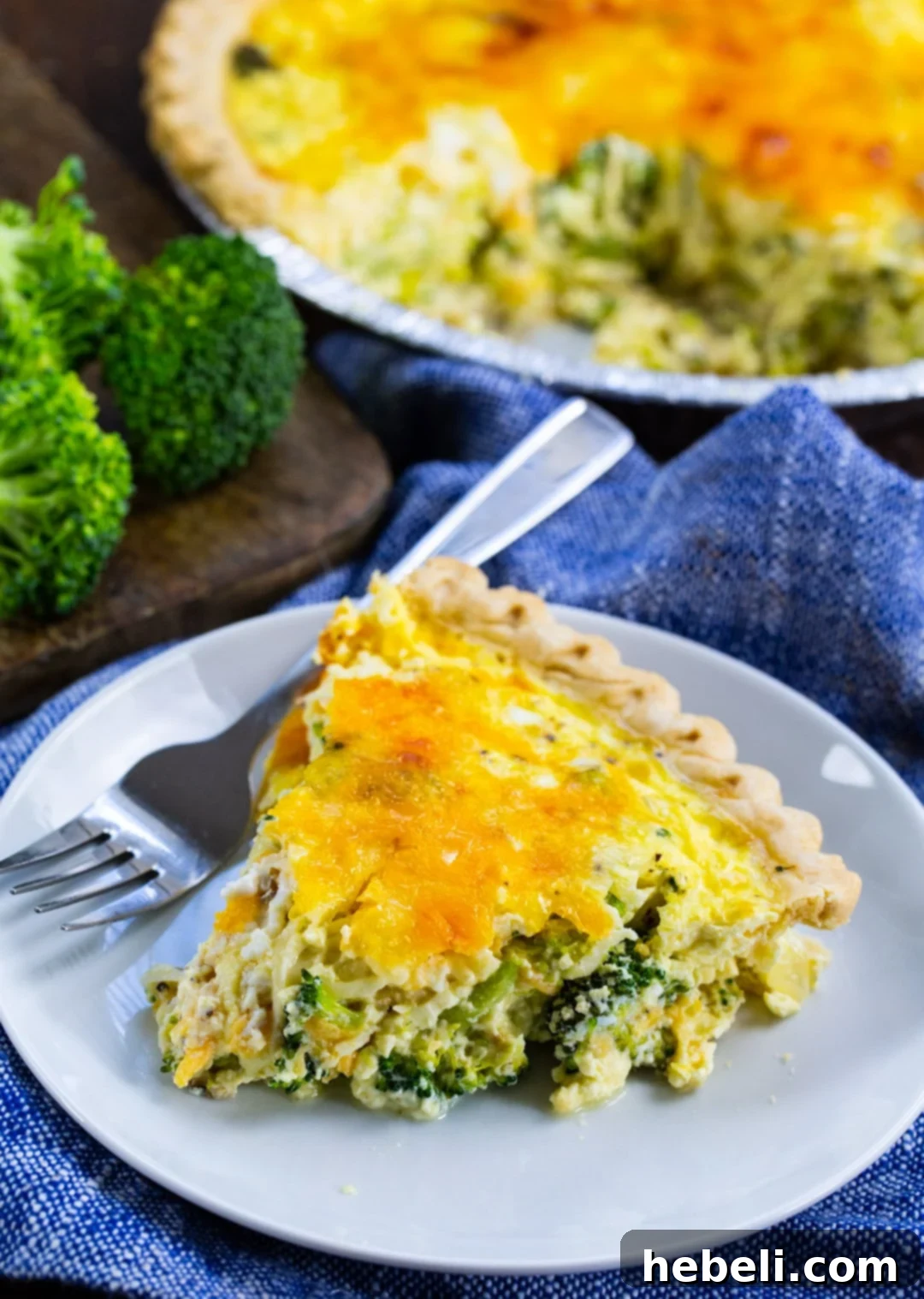 A generous, golden-crusted slice of Broccoli Quiche resting on a plate, with the remainder of the beautifully baked quiche visible in the soft-focus background, inviting you to enjoy its rich flavors.