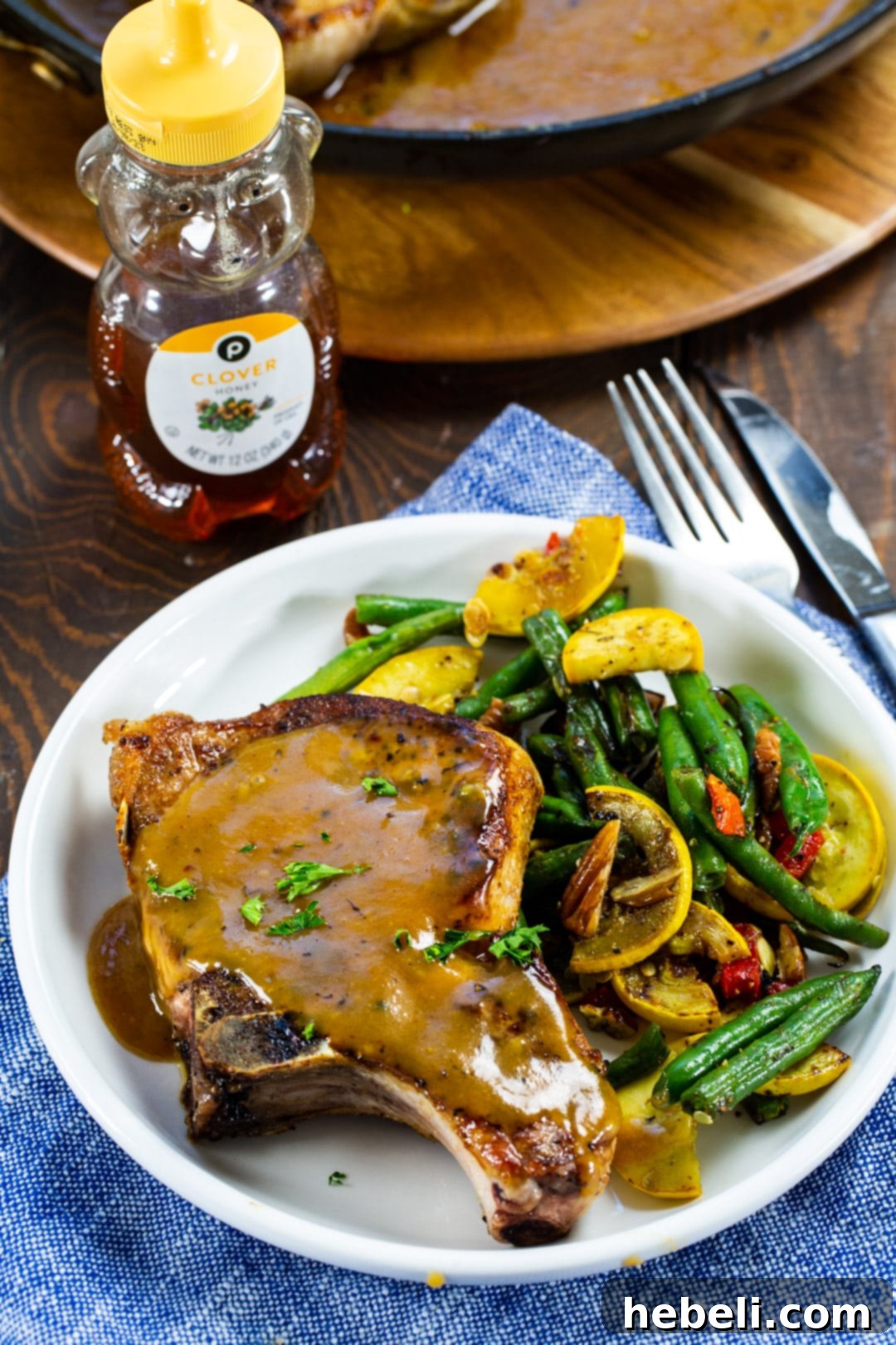 Glazed Honey Mustard Pork Chops 3 Close-up of a Honey Mustard Pork Chop on a plate, garnished with green beans and squash, with a bottle of honey in the background.