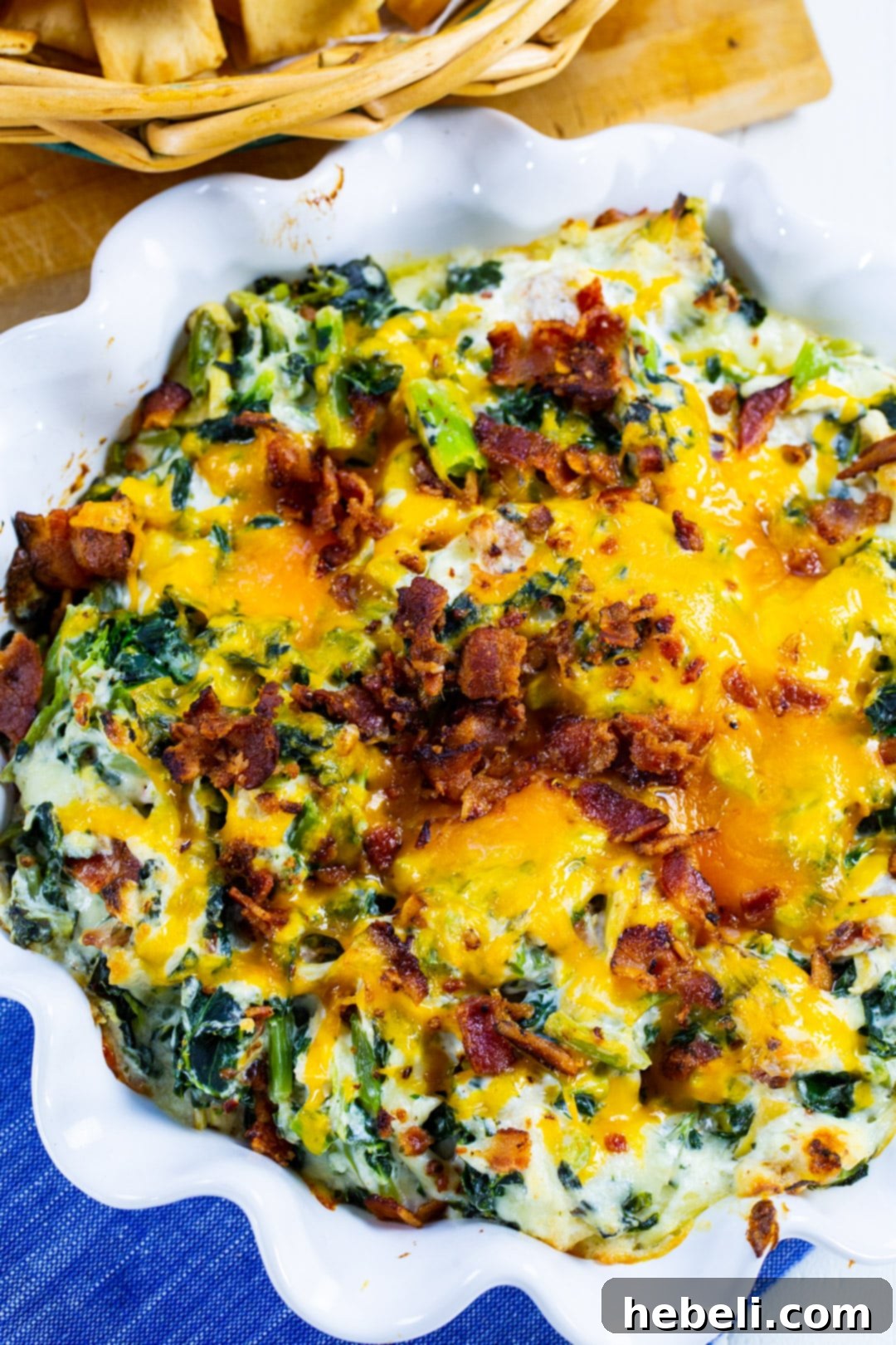 A close-up view of the golden-baked Collards Artichoke Dip with visible melted cheese and greens, served in an elegant round fluted baking dish, garnished with fresh herbs.
