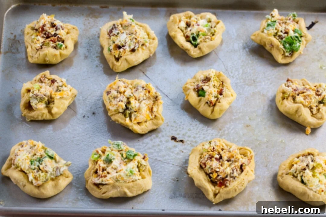 Unbaked biscuit rounds neatly arranged on a baking sheet, each topped with creamy filling and a jalapeno slice, ready for the oven.
