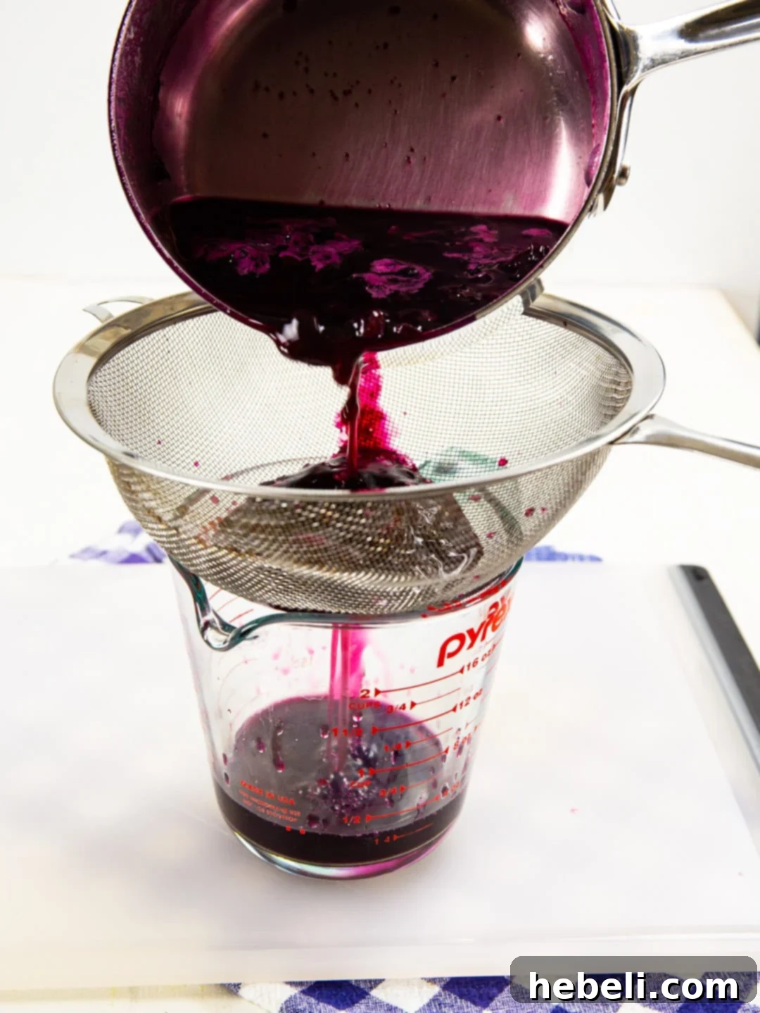 Vibrant purple blueberry simple syrup being strained through a fine-meshed sieve into a glass measuring cup, separating the liquid from the fruit solids.