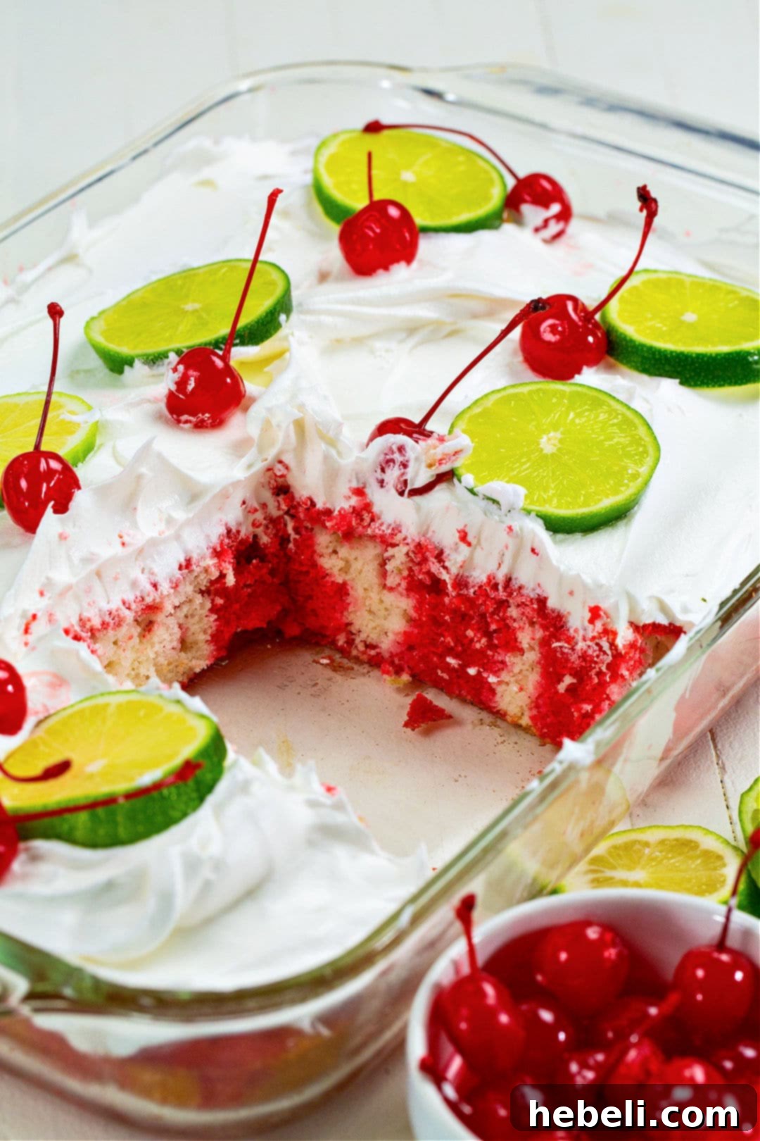 Overhead view of a fully assembled Cherry Limeade Poke Cake in its baking dish, topped with Cool Whip, lime slices, and cherries.