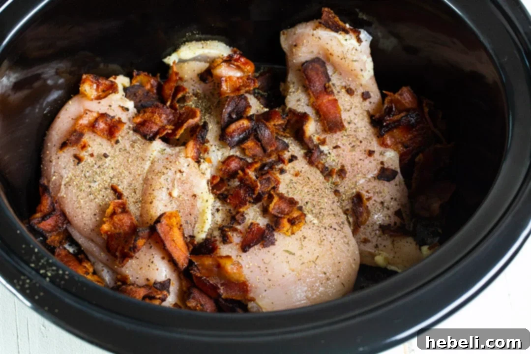 Uncooked boneless, skinless chicken breasts seasoned with spices and topped with crumbled bacon in a slow cooker, ready for the gravy mixture.