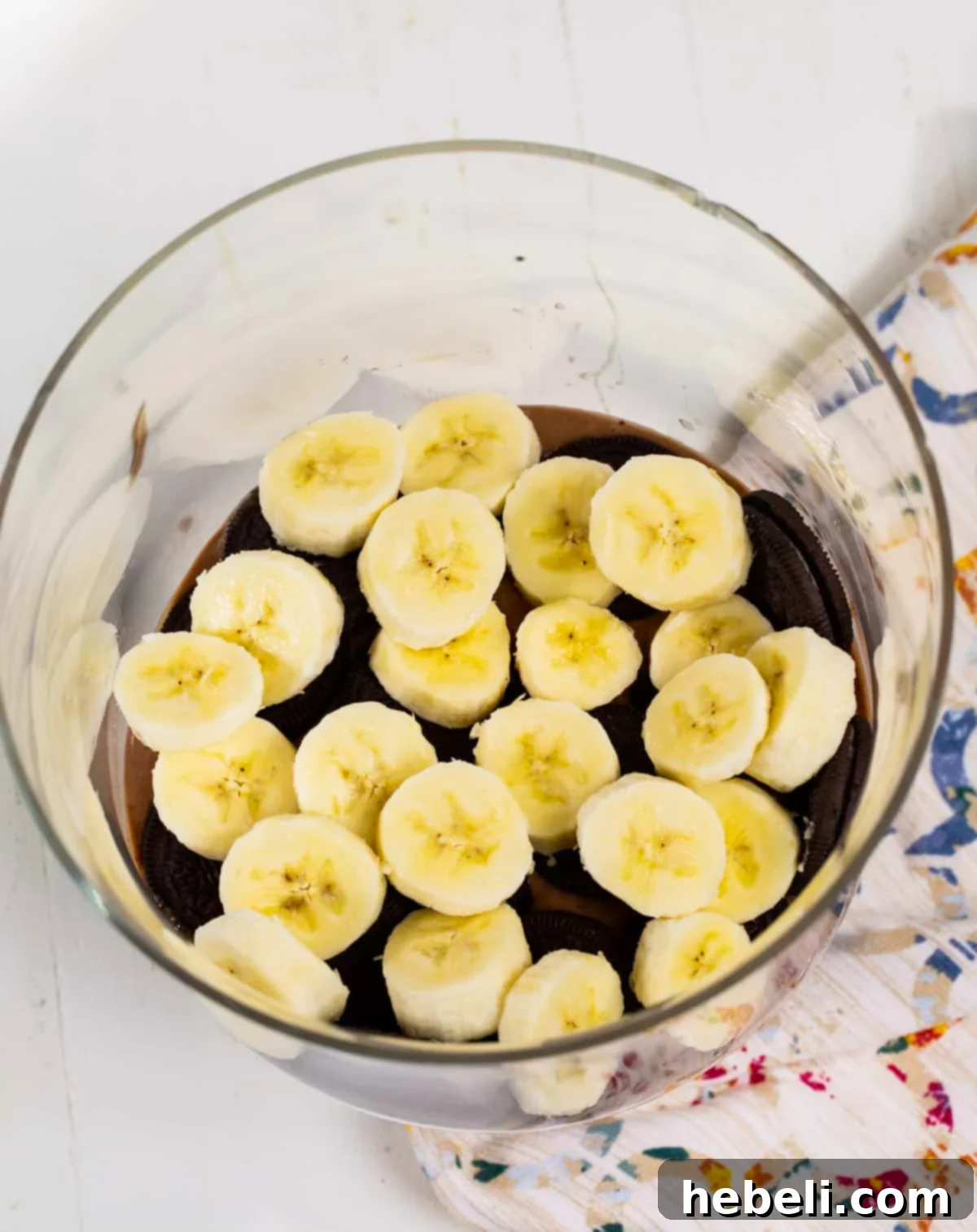 Decadent Nutella Banana Pudding 4 Freshly sliced banana pieces are carefully arranged over a layer of Oreo cookies within a trifle dish, preparing for the next layer of creamy Nutella pudding.