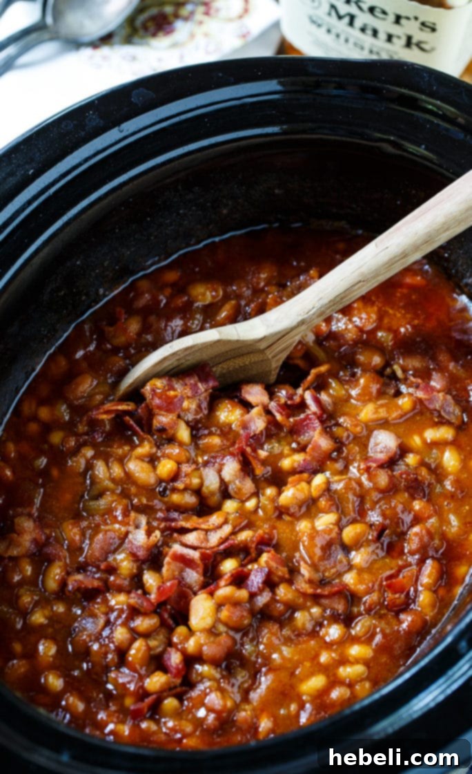 A close-up of Slow Cooker Bourbon Baked Beans in a black slow cooker, showing the rich, thick sauce and bacon pieces.