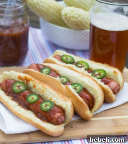 Signature Dr. Pepper BBQ Hot Dogs 3 Hot Dogs on a wooden cutting board with a cold beer and corn in the background, showing a relaxed barbecue setting.