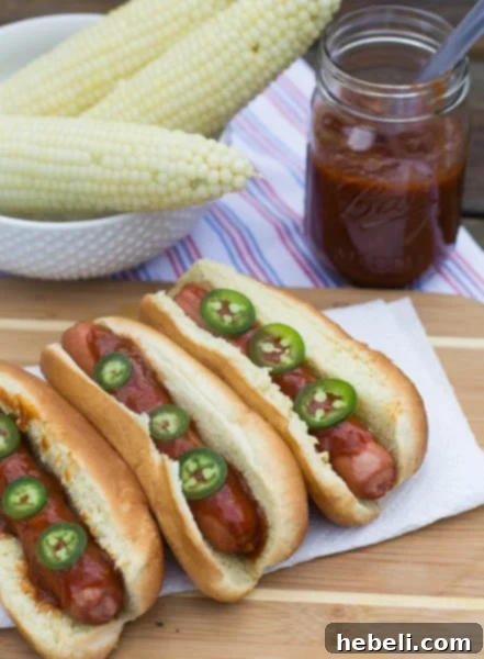 Signature Dr. Pepper BBQ Hot Dogs 4 Three hot dogs topped with Dr. Pepper Barbecue Sauce and fresh jalapeno slices, with corn on the cob in the background.