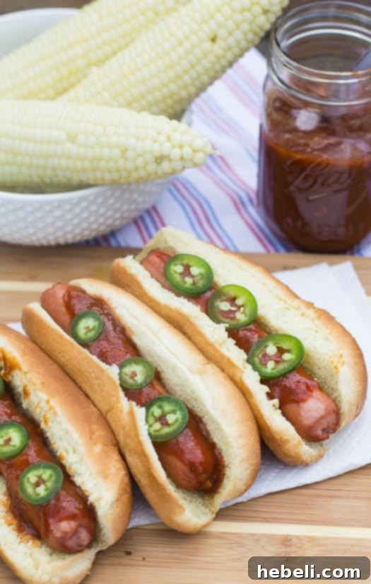 Signature Dr. Pepper BBQ Hot Dogs 6 A vibrant image of hot dogs generously coated with Dr. Pepper BBQ Sauce, served with grilled corn on the cob in the background.