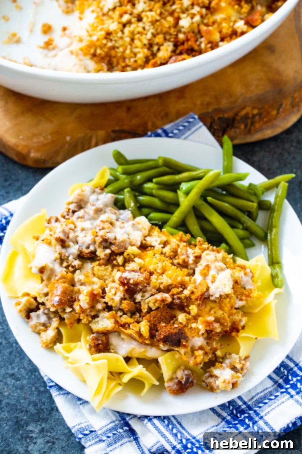 A close-up shot of Sandy's Chicken, featuring the golden-brown crust of the stuffing and melted cheese, served with a generous portion of egg noodles and crisp green beans on a white plate.